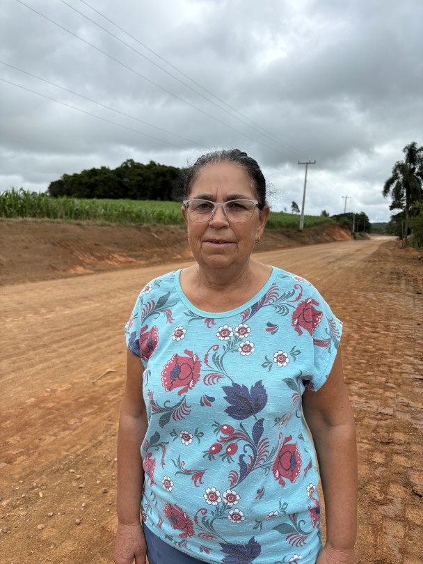 A imagem apresenta uma mulher de meia-idade, com pele clara e cabelos escuros parcialmente presos, usando óculos de armação transparente. Ela veste uma camiseta de manga curta azul-clara com estampas florais vibrantes em tons de vermelho, branco e azul-escuro. A mulher está em pé, posicionada do peito para cima, olhando diretamente para a câmera com uma expressão neutra. Ela se encontra em um ambiente externo, em uma estrada de terra batida de cor avermelhada, que se estende ao fundo. No lado esquerdo da imagem, é possível ver um campo de cultivo verde, provavelmente milho, e algumas árvores distantes. No lado direito, há mais vegetação, um poste de energia elétrica e fios que cruzam o céu nublado. A iluminação é difusa, indicando um dia nublado.