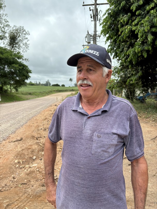A imagem mostra um homem de meia-idade, com pele clara, cabelos grisalhos visíveis nas laterais e um bigode branco. Ele está usando um boné escuro com a aba virada para trás, que tem a palavra "COMIL" parcialmente visível em cores vibrantes, e uma camiseta polo de cor azul acinzentada com um bolso no peito. Ele está posicionado em um ângulo de três quartos, olhando para o lado direito da imagem com uma expressão pensativa. O cenário atrás dele é uma estrada de terra batida de cor avermelhada. No lado esquerdo da estrada, há um campo verde com algumas árvores ao fundo. No lado direito, próximo ao homem, há um poste de energia elétrica com vários fios e um emaranhado de cabos, e algumas árvores com folhagem densa. O céu está nublado, indicando um dia com pouca luz solar direta.