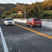 A imagem mostra uma ponte com dois carros trafegando em sentidos opostos. A estrutura possui barreiras brancas nas laterais e marcação central amarela. Ao fundo, há uma paisagem rural com colinas, árvores e algumas casas.