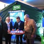 A imagem mostra quatro pessoas em pé ao redor de uma pequena mesa redonda, com uma pessoa assinando um documento. Eles estão em frente a uma tenda verde com o logotipo "SESI". Também há caixas azuis com o texto "Sistema Fiergs" visíveis ao fundo.
