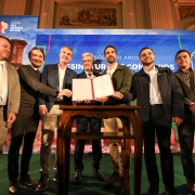 A imagem mostra um grupo de oito homens em um evento formal, com um palco e um telão ao fundo.

No centro da imagem, três homens estão em frente a uma pequena mesa de madeira escura, sobre a qual repousam dois documentos em pastas vermelhas abertas.

O homem mais ao centro, de cabelo grisalho e terno azul escuro, está com as mãos sobre os documentos, parecendo assinar ou apresentar algo.
À sua esquerda (direita da imagem), um homem de camisa azul clara e blazer cinza sorri e aponta para os documentos.
À sua direita (esquerda da imagem), um homem de camisa verde-oliva e blazer cinza também tem uma das mãos sobre os documentos.
Os outros cinco homens estão posicionados ao redor. Da esquerda para a direita:

O primeiro homem, na extrema esquerda, veste um terno claro e calças que parecem de montaria.
O segundo homem usa terno escuro e gravata.
Os dois homens mais à direita, também de terno, estão com os braços cruzados ou as mãos unidas.
O último homem, na extrema direita, tem barba e está com os braços cruzados.
Ao fundo, há um grande telão com gráficos coloridos e a frase "SÕES 400 ANOS". À esquerda do telão, pode-se ver um logotipo com a inscrição "GOVERNO RIO GRANDE DO SUL". O ambiente é bem iluminado e parece ser um salão de eventos.