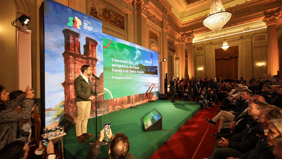 A imagem retrata um evento formal em um grande salão, onde um homem está discursando em um palco e uma plateia o assiste.
No centro da imagem, em um palco elevado coberto por um carpete verde, um homem de barba, vestindo uma jaqueta verde-oliva, camisa branca e calças cáqui, está de pé em frente a um microfone. Ele parece estar apresentando algo, talvez utilizando um controle remoto na mão direita.
Atrás dele, há um grande telão dividido em duas seções. A seção da esquerda exibe uma imagem de ruínas históricas e, no canto superior esquerdo, um logotipo com "GOVERNO RIO GRANDE DO SUL". A seção da direita mostra um texto que diz "Concessão dos aeroportos de Passo Fundo e de Santo Ângelo" e abaixo "Projeto revisado".
À direita do palco, há uma grande plateia sentada em cadeiras, com muitas pessoas observando o orador. Algumas pessoas, no lado esquerdo da imagem, estão em pé e tirando fotos com seus celulares.
O salão é grandioso, com teto alto, paredes ornamentadas, colunas e lustres de cristal suspensos, sugerindo um ambiente histórico ou governamental. O piso à frente do palco é vermelho e há iluminação focada no palco.