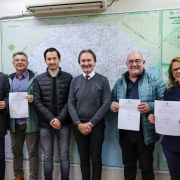 Na imagem aparecem seis pessoas sorridentes, cinco homens e uma mulher. Eles posam para uma foto em frente a um grande mapa rodoviário. As duas pessoas do lado esquerdo da foto e as duas que estão do lado direito seguram papéis. A mulher à direita usa óculos e um casaco azul-escuro.