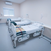 Imagem mostra um quarto de hospital claro e moderno com três camas brancas arrumadas. Há um ar condicionado na parede superior e uma janela com persianas.