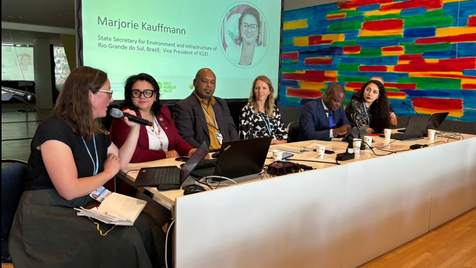 A foto mostra seis pessoas sentadas lado a lado atrás de uma mesa comprida, com notebooks e microfones à frente. Atrás delas, há uma tela grande com o nome "Marjorie Kauffmann" e o título "Secretária Estadual do Meio Ambiente e Infraestrutura do Rio Grande do Sul; Vice-presidente do ICLEI". A parede ao fundo tem uma arte abstrata colorida.