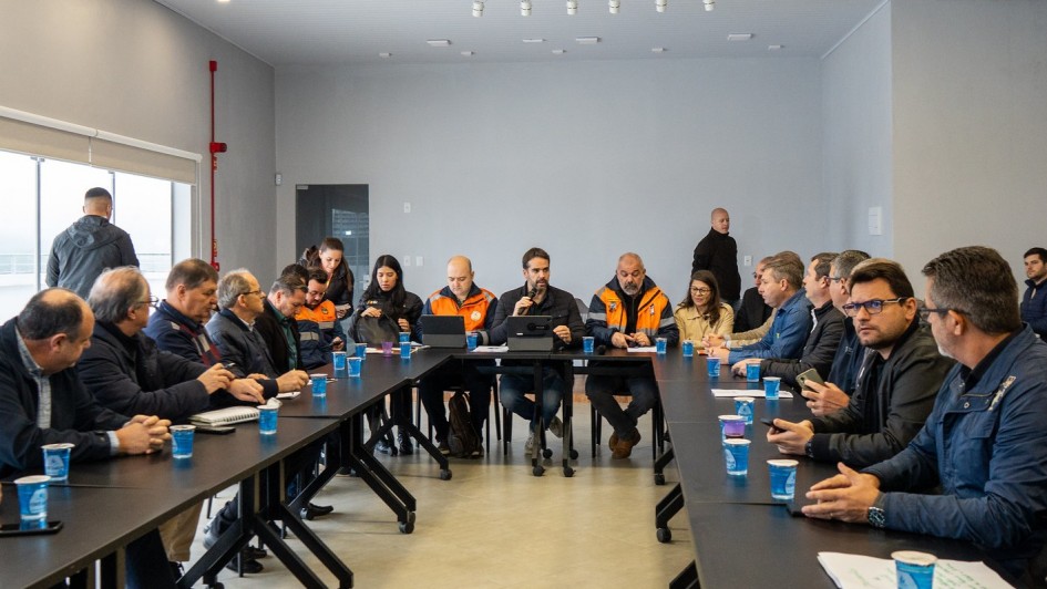 A imagem mostra uma reuni&atilde;o formal com v&aacute;rias pessoas sentadas ao redor de uma mesa, algumas usando jaquetas laranja, possivelmente representando uma equipe ou &oacute;rg&atilde;o espec&iacute;fico.