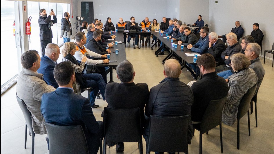 A foto mostra uma sala de reuniões com uma grande mesa em formato de U, ao redor da qual estão sentadas cerca de 25 pessoas. Algumas estão de pé, tirando fotos ou gravando vídeos. Sobre as mesas, há copos e papéis espalhados. Um dos lados da sala tem janelas grandes, por onde entra bastante luz natural, iluminando o ambiente.