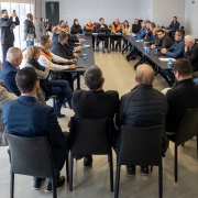 A foto mostra uma sala de reuniões com uma grande mesa em formato de U, ao redor da qual estão sentadas cerca de 25 pessoas. Algumas estão de pé, tirando fotos ou gravando vídeos. Sobre as mesas, há copos e papéis espalhados. Um dos lados da sala tem janelas grandes, por onde entra bastante luz natural, iluminando o ambiente.