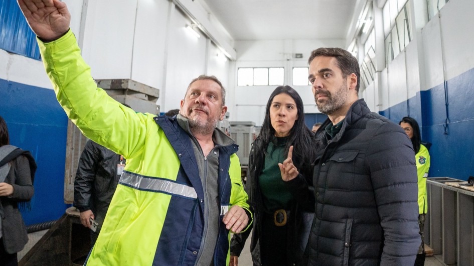 A imagem mostra uma visita a uma casa de bombeamento de água. À direita está o governador Eduardo Leite, de casaco preto, observando atentamente enquanto um homem com casaco amarelo com faixas refletivas aponta algo fora da imagem. Ao centro, uma mulher com casaco preto acompanha a cena.