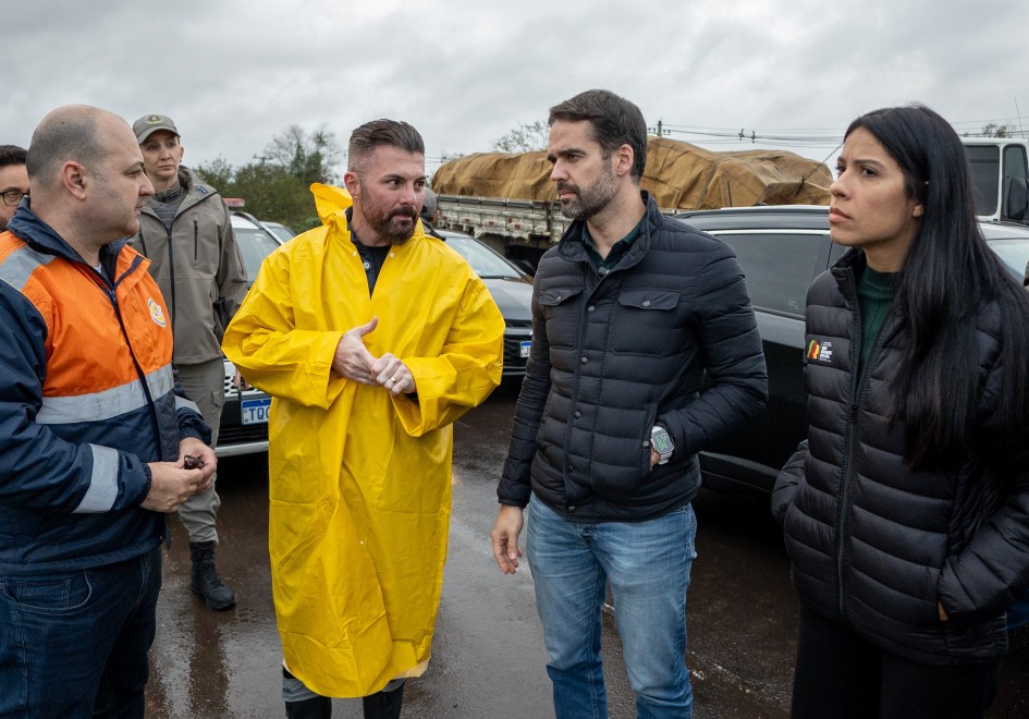 A imagem mostra o governador Eduardo Leite, de jaqueta preta, em um estacionamento ao ar livre, acompanhado por outras pessoas com roupas de chuva e jaquetas coloridas, em um cenário que sugere visita a uma área atingida por fortes chuvas. Ao fundo, há caminhões e veículos estacionados.