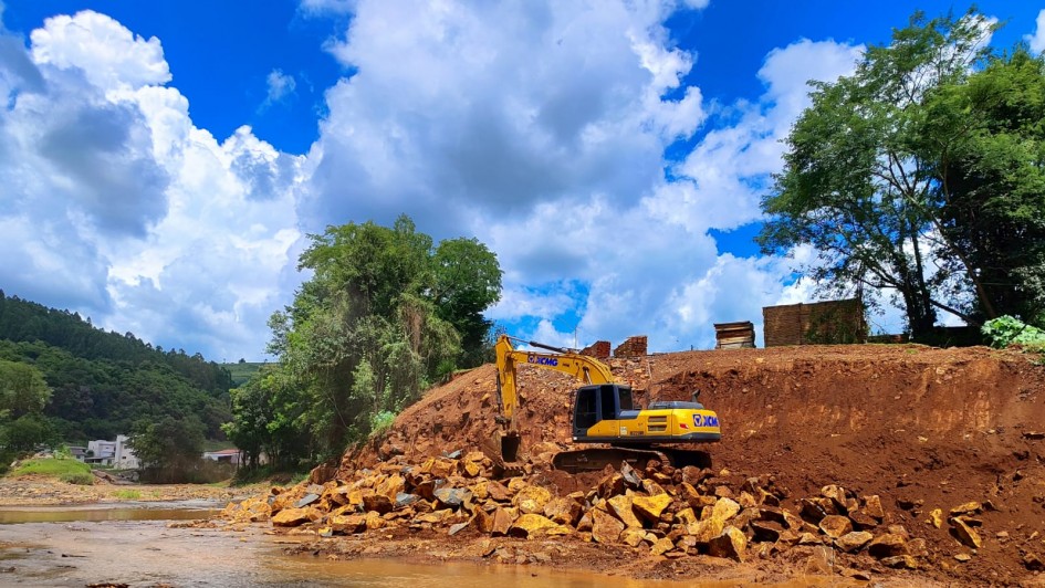 A imagem mostra uma escavadeira amarela operando em um terreno com pedras e terra, em meio a uma área com vegetação e construções ao fundo. A cena representa trabalhos de desassoreamento realizados no município de Barra do Rio Azul, voltados à prevenção de enchentes.