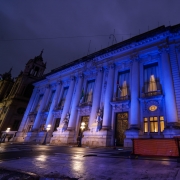 A imagem mostra o Palácio Piratini, sede do governo do Rio Grande do Sul, iluminado com luz azul durante a noite. A fachada neoclássica com colunas e esculturas está em destaque. A iluminação azul faz referência ao Dia Mundial do Refugiado.