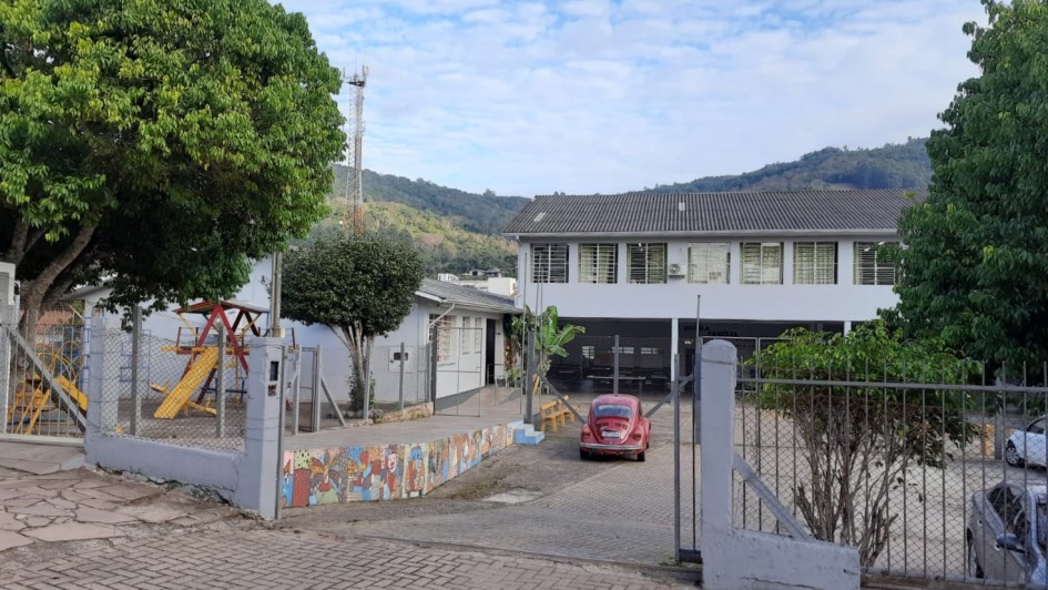 Imagem mostra portão de acesso a pátio de escola. Ao fundo, prédio de dois andares com janelas de vidros no segundo andar. Na esquerda da imagem aparece uma pracinha de brinquedos.