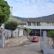 Imagem mostra portão de acesso a pátio de escola. Ao fundo, prédio de dois andares com janelas de vidros no segundo andar. Na esquerda da imagem aparece uma pracinha de brinquedos.