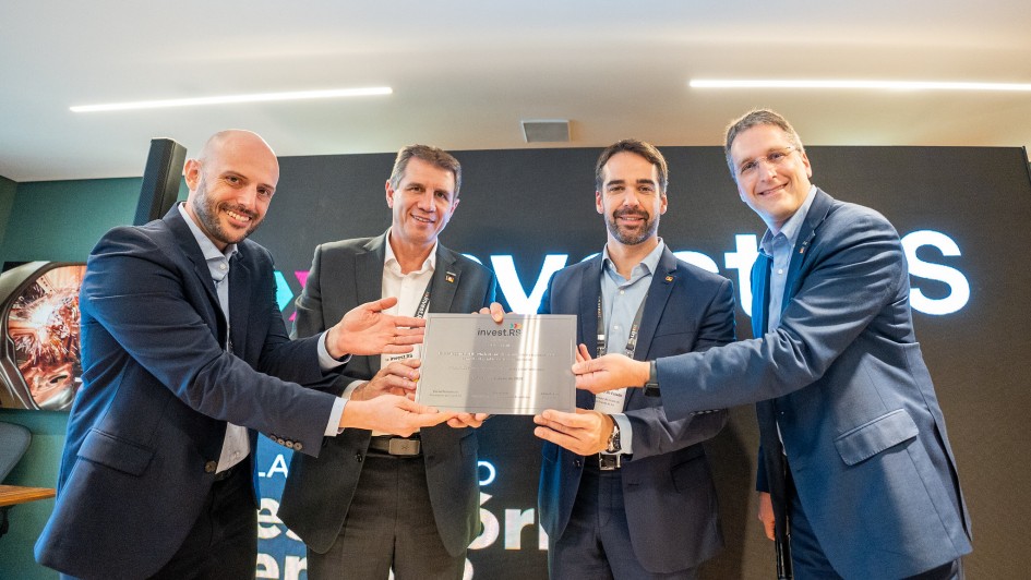 Na imagem, Eduardo Leite é a figura central segurando um documento ou placa ao lado de outras três pessoas, todos vestidos formalmente. O ambiente sugere um evento institucional da Invest RS, com um telão ao fundo reforçando o caráter oficial da ocasião.