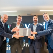 Na imagem, Eduardo Leite é a figura central segurando um documento ou placa ao lado de outras três pessoas, todos vestidos formalmente. O ambiente sugere um evento institucional da Invest RS, com um telão ao fundo reforçando o caráter oficial da ocasião.