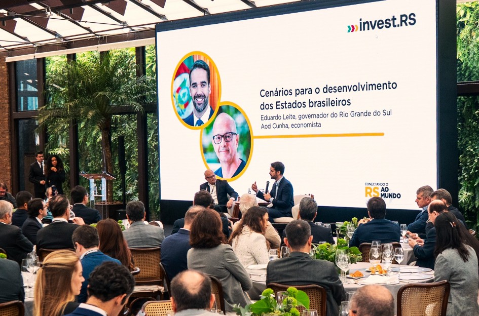 A imagem mostra Eduardo Leite, governador do Rio Grande do Sul, discursando ao microfone em um evento formal. Ao fundo, um telão exibe o tema “Cenários para o desenvolvimento dos Estados brasileiros”, indicando uma palestra ou debate promovido pela Invest RS.