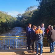 A imagem mostra um grupo de pessoas em pé sobre uma ponte sobre um rio, cercado por árvores e vegetação. Há também um tripé vermelho com equipamentos montados no lado esquerdo da imagem.