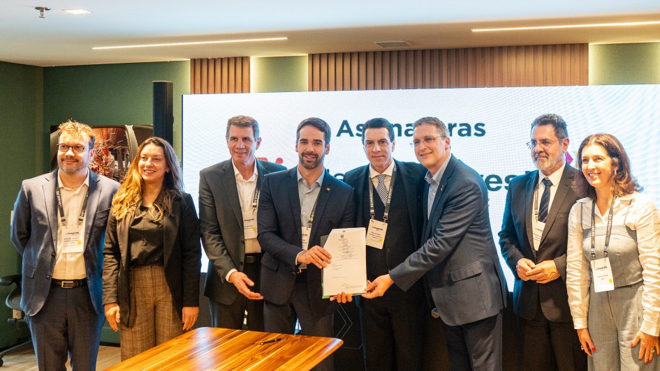 A imagem mostra um grupo de oito pessoas reunidas em um ambiente formal, com algumas segurando um documento. Ao fundo, há uma grande tela exibindo parcialmente os textos "Assinaturas" e "RS". Há também uma mesa de madeira em primeiro plano.