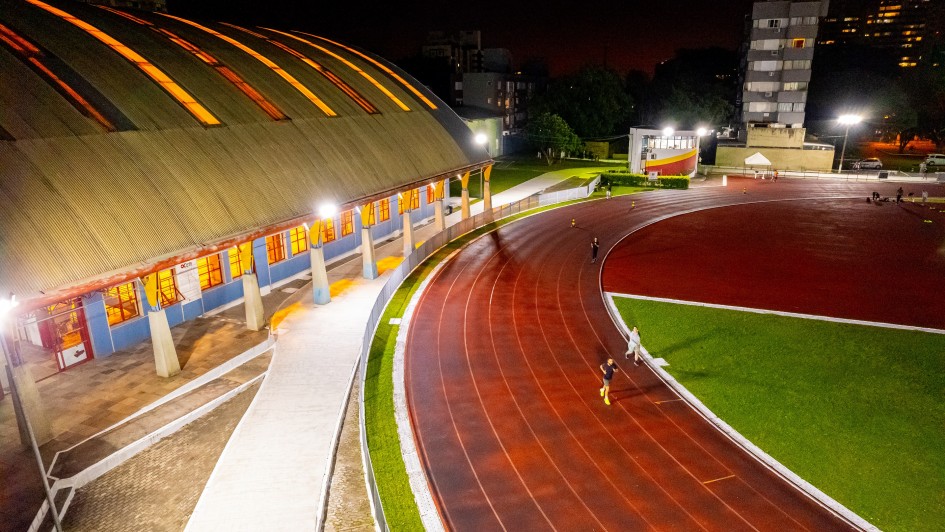 A foto noturna em alta definição mostra a pista de atletismo e o ginásio do Centro Estadual de Treinamento Esportivo (Cete) iluminados. A pista de corrida é vermelha escura, com algumas raias visíveis, e a área central é um campo de grama verde vibrante. Algumas pessoas, em tamanho pequeno devido à distância, estão correndo na pista. À esquerda, destaca-se a fachada de um grande ginásio com um telhado abobadado em tons de dourado e laranja devido à iluminação interna. As paredes do ginásio são azuis na parte inferior e brancas nos pilares. Postes de luz altos e brilhantes iluminam intensamente toda a área. Ao fundo, edifícios e árvores são visíveis sob o céu escuro.