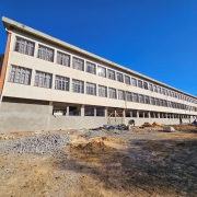 A imagem mostra um prédio de três andares em construção. A estrutura tem formato retangular e alongado, com várias janelas alinhadas horizontalmente em cada andar. O prédio está localizado em um terreno de terra batida, e ao redor dele há pilhas de materiais de construção espalhadas, indicando que a obra ainda está em andamento. O céu está claro e azul, com boa iluminação natural.