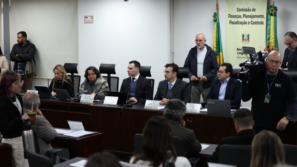 A imagem retrata um ambiente formal de reunião ou conferência. Várias pessoas estão sentadas em uma longa mesa voltada para o público. Sobre a mesa, há placas com nomes e laptops. Ao fundo, um banner exibe o texto "Comissão de Finanças, Planejamento, Fiscalização e Controle", acompanhado de algumas bandeiras. Algumas pessoas estão em pé observando, e uma delas parece estar operando uma câmera.