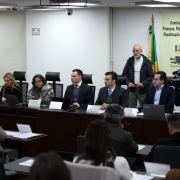 A imagem retrata um ambiente formal de reunião ou conferência. Várias pessoas estão sentadas em uma longa mesa voltada para o público. Sobre a mesa, há placas com nomes e laptops. Ao fundo, um banner exibe o texto "Comissão de Finanças, Planejamento, Fiscalização e Controle", acompanhado de algumas bandeiras. Algumas pessoas estão em pé observando, e uma delas parece estar operando uma câmera.