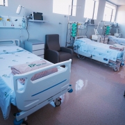 A imagem mostra um quarto de hospital com três leitos equipados. Cada leito possui: uma cama hospitalar ajustável, coberta com lençóis brancos que exibem o logotipo do hospital; equipamentos médicos ao lado, como monitores e bombas de infusão, e cadeiras reclináveis próximas às camas, provavelmente destinadas a acompanhantes ou visitantes. Ao fundo, há janelas que permitem a entrada de luz natural, iluminando o ambiente. No canto direito da imagem, aparece uma pessoa vestida com roupas verdes, típicas de profissionais da saúde.
