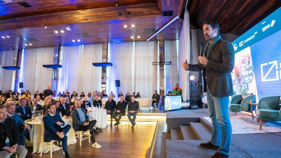 Na imagem, o governador Eduardo Leite aparece discursando em um evento realizado em um salão amplo e bem iluminado, com piso e teto de madeira. Ele está de pé, segurando um microfone, e se dirige a uma plateia atenta, sentada em mesas redondas. O ambiente é elegante, com iluminação azul e janelas altas com cortinas brancas. Ao fundo, há um telão exibindo logotipos, incluindo o da Corsan, sinalizando a formalidade e importância institucional do encontro.