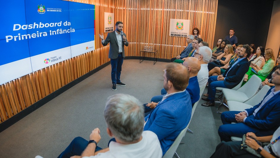 A foto mostra uma apresentação em um auditório. No centro, o vice-governador e coordenador do Ceipi, Gabriel Souza, um homem de barba, terno escuro e camisa clara, está de pé no palco. Ele segura um microfone e gesticula com a mão esquerda em direção a um telão azul à sua esquerda, que exibe "Dashboard da Primeira Infância" e o logotipo do Governo do Estado do Rio Grande do Sul. O palco tem um painel de madeira com listras verticais. Uma plateia diversificada, composta por homens e mulheres, está sentada em cadeiras claras, atenta à apresentação. No fundo, há outro painel com o logotipo do governo.