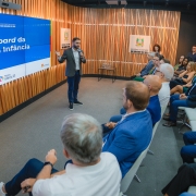 A foto mostra uma apresentação em um auditório. No centro, o vice-governador e coordenador do Ceipi, Gabriel Souza, um homem de barba, terno escuro e camisa clara, está de pé no palco. Ele segura um microfone e gesticula com a mão esquerda em direção a um telão azul à sua esquerda, que exibe "Dashboard da Primeira Infância" e o logotipo do Governo do Estado do Rio Grande do Sul. O palco tem um painel de madeira com listras verticais. Uma plateia diversificada, composta por homens e mulheres, está sentada em cadeiras claras, atenta à apresentação. No fundo, há outro painel com o logotipo do governo.
