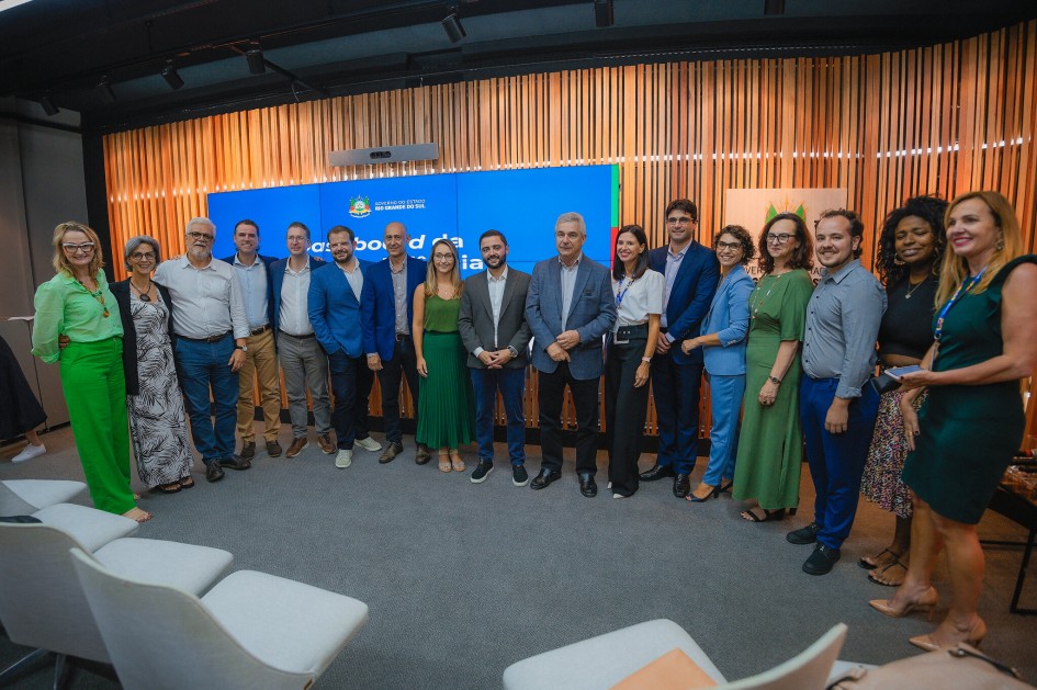A foto mostra um grupo de cerca de 18 pessoas, incluindo homens e mulheres, de diversas idades, posando juntas para uma foto em um auditório moderno. Eles estão em pé, sorridentes, formando um semicírculo em frente a um palco. Ao fundo, um telão azul com o logotipo do Governo do Estado do Rio Grande do Sul e a palavra "Dashboard" parcialmente visível, além de um painel de madeira com listras verticais. A maioria das pessoas está vestindo roupas sociais ou esporte fino, em tons variados. Há um contraste de cores, com algumas pessoas em tons vibrantes de verde e azul. No primeiro plano, algumas cadeiras vazias em tons de branco e cinza.