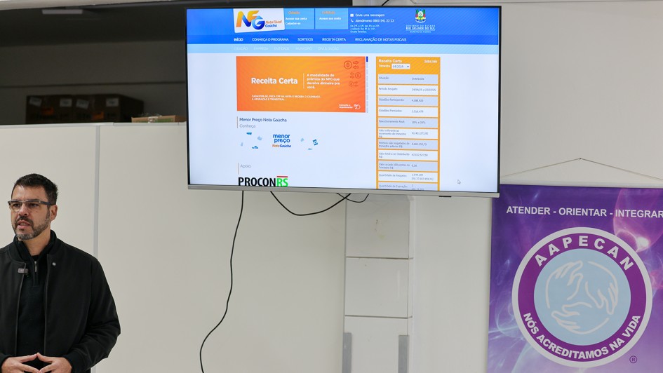 A imagem mostra uma pessoa em pé ao lado de uma mesa, sobre a qual há um laptop aberto. Acima da pessoa, há uma tela grande exibindo um site do programa Nota Fiscal Gaúcha. À direita da imagem, há um banner vertical da Aapecan (Associação de Apoio a Pessoas com Câncer). O banner contém o lema "ATENDER • ORIENTAR • INTEGRAR" e um emblema com mãos dadas dentro de um círculo, simbolizando apoio e solidariedade.