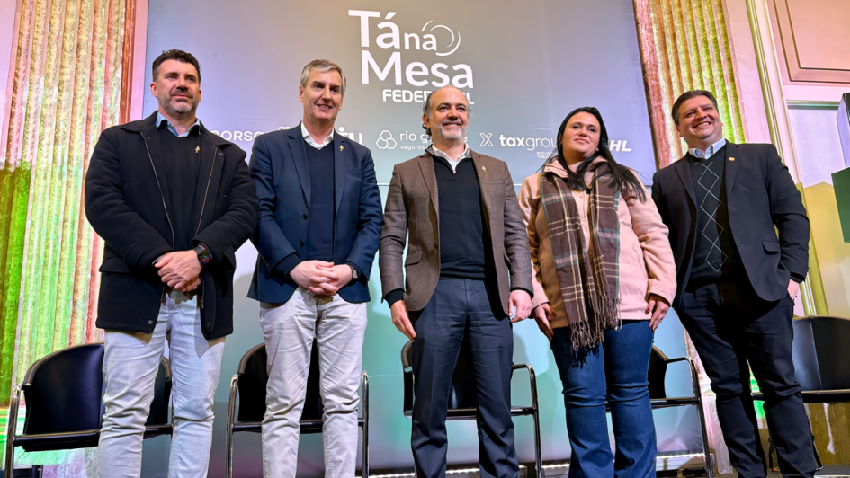 Foto do encontro “Tá na Mesa", promovido pela Federasul, na qual o secretário de Estado da Cultura, Eduardo Loureiro posa, ao lado de de outras quatro pessoas, no palco do evento. Ao fundo, há um painel com o título “Tá na Mesa – Federasul” e logotipos de patrocinadores. Todos estão vestidos de forma formal ou casual elegante, com casacos, blazers e cachecol, sugerindo clima frio. Atrás deles, há cadeiras vazias, além de iluminação em tons de verde e rosa. O ambiente é um salão de eventos.