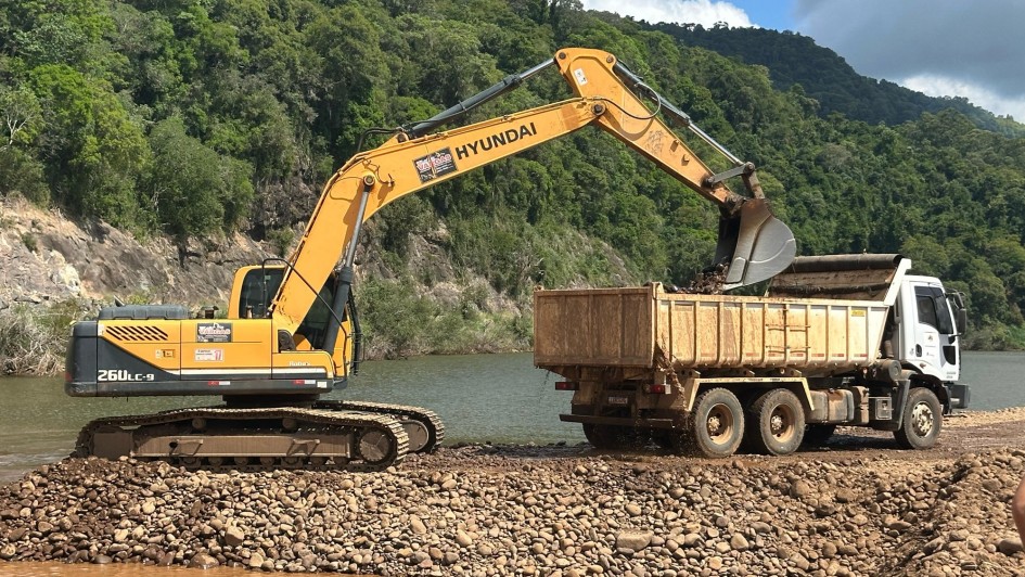 A imagem mostra uma escavadeira Hyundai 260LC-9 carregando pedras em um caminhão basculante, em uma área próxima a um rio. O solo é composto por pedras e cascalho, e ao fundo há vegetação densa. A cena representa uma etapa de desassoreamento ou intervenção de infraestrutura para controle de enchentes ou recuperação ambiental.