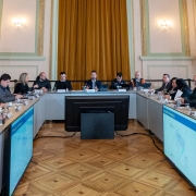 A imagem mostra uma sala de conferências com várias pessoas sentadas ao redor de uma mesa em formato de U, com o governador ao centro. Em frente a cada participante, há plaquinhas com nomes, microfones e documentos, sugerindo uma reunião formal ou institucional. Em ambos os lados da mesa, há duas telas grandes exibindo informações meteorológicas.