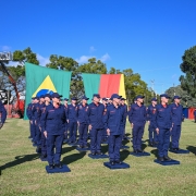 A imagem retrata uma cerimônia de formatura do Corpo de Bombeiros Militar do Rio Grande do Sul. Bombeiros uniformizados estão perfilados em um gramado, com bandeiras do Brasil e do RS ao fundo, além de um caminhão e equipamentos de resgate visíveis. O cenário é ao ar livre, com céu limpo e árvores ao redor, transmitindo um clima solene e de celebração da conquista profissional.