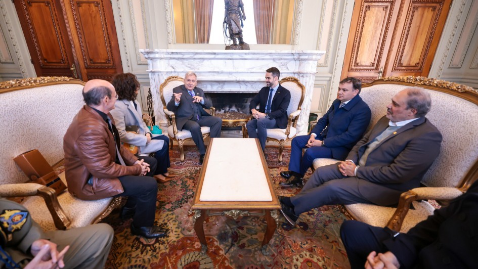 Foto mostra um elegante salão com lareira e móveis clássicos, um grupo de homens e uma mulher sentam-se em cadeiras estofadas, engajados em uma conversa. Dois homens, aparentemente em destaque, estão sentados mais próximos à lareira.