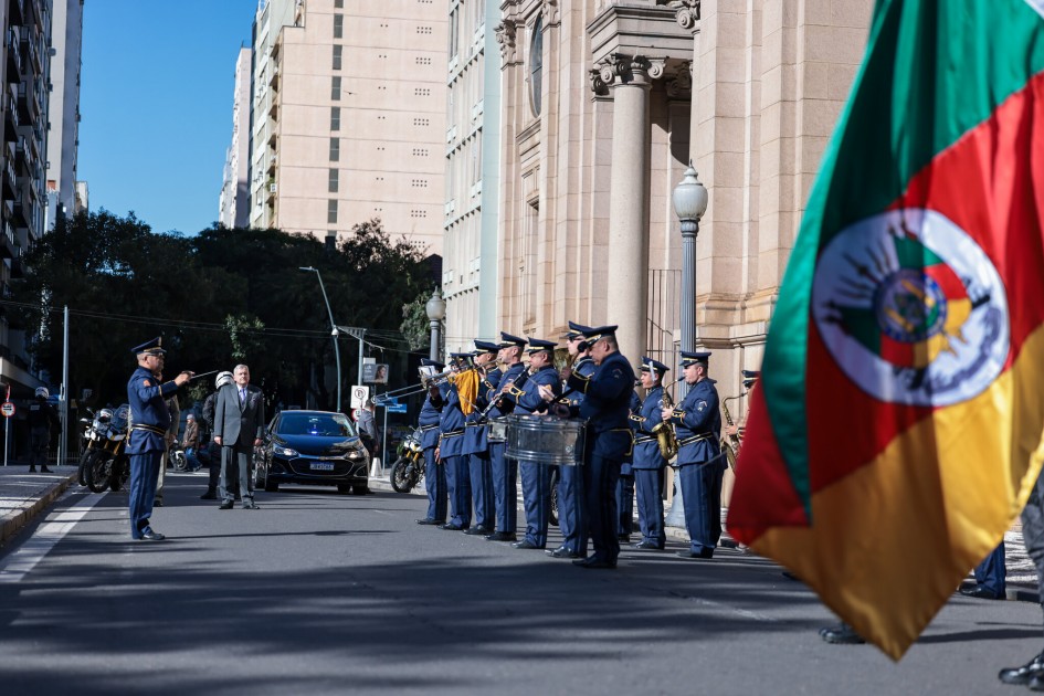 Uma banda militar em uniforme azul, alinhada na rua, toca instrumentos enquanto um carro escuro se aproxima em frente a um edifício clássico. No canto direito, a bandeira do Rio Grande do Sul em primeiro plano, com suas cores vibrantes, tremula ao vento.