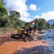 A imagem mostra uma escavadeira operando na margem de um rio. A máquina está removendo sedimentos da beira do rio, o que faz com que a água próxima fique turbulenta e barrenta. Ao redor, a paisagem é rica em vegetação verde, com árvores densas e mato cobrindo o terreno. 