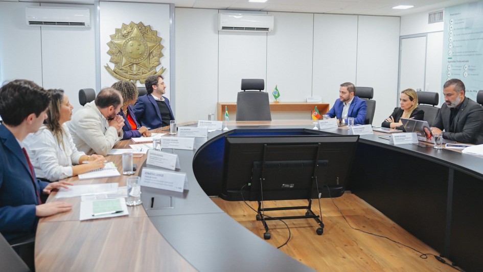 A imagem mostra o vice-governador Gabriel Souza em uma reunião oficial em Brasília, em uma sala de conferências com mesa oval, bandeiras do Brasil e do Rio Grande do Sul ao fundo e o brasão da República na parede. A cena reflete um ambiente institucional, onde Gabriel articula apoio para a construção de pontes internacionais entre o RS e a Argentina.