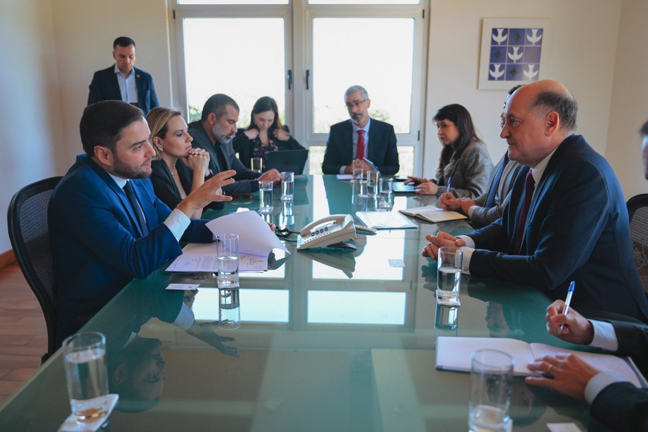 A imagem mostra o vice-governador Gabriel Souza em uma reunião formal em Brasília, ao redor de uma mesa de vidro com outros participantes. O ambiente é iluminado e profissional, com documentos e copos d’água sobre a mesa, indicando tratativas sérias relacionadas à articulação de apoio para projetos de infraestrutura entre o Rio Grande do Sul e a Argentina.