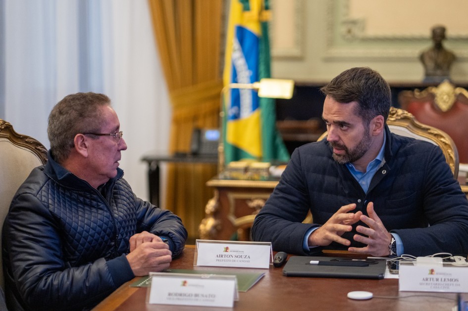 A imagem mostra o governador Eduardo Leite em diálogo direto com o prefeito de Canoas, Airton Souza, durante uma reunião formal. Ambos estão sentados à mesa, acompanhados por outros participantes identificados por placas nominais. O ambiente é institucional, com bandeira do Brasil ao fundo e cortinas douradas, sugerindo o Palácio Piratini como local do encontro. A cena transmite um momento de articulação política e administrativa, possivelmente relacionado à liberação de recursos para obras de contenção de cheias no município.