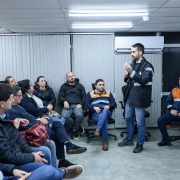 A foto mostra o governador Eduardo Leite (ao centro, em pé, de casaco escuro) conversando com um grupo de pessoas sentadas em uma sala. Ele segura um objeto pequeno em uma das mãos enquanto gesticula com a outra, parecendo explicar algo. As pessoas sentadas, algumas vestindo coletes da Defesa Civil, o observam atentamente. A sala parece ser um ambiente de escritório ou reunião, com persianas e ar condicionado. No canto superior direito, há um monitor exibindo parte de um texto.