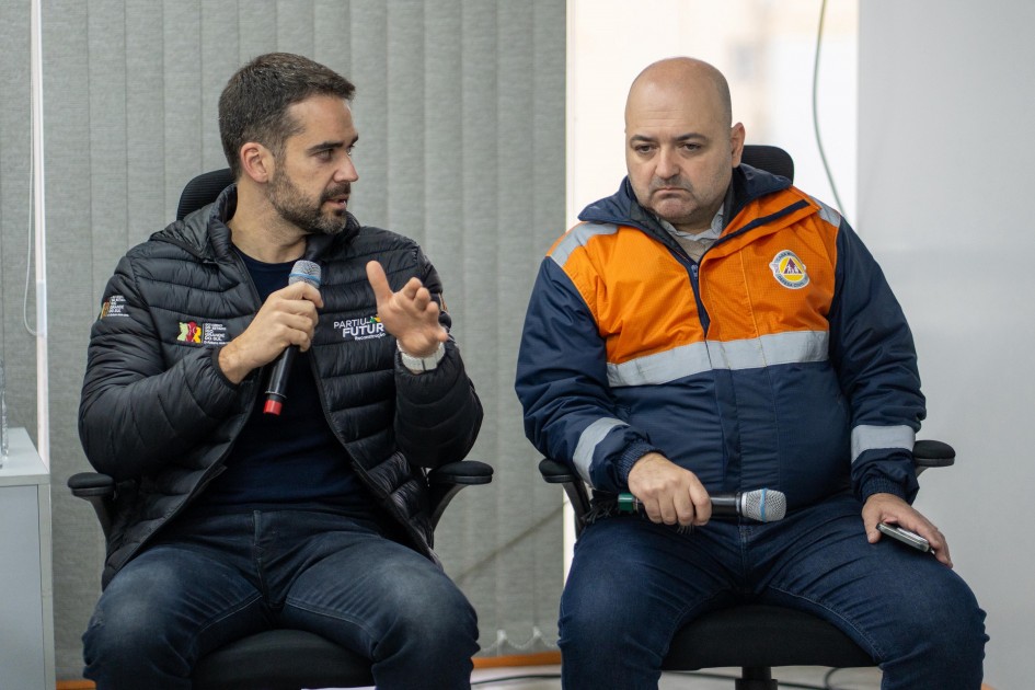 A foto mostra o governador Eduardo Leite e o Cel. Luciano Chaves Boeira em uma reunião. Eduardo Leite, à esquerda, de casaco preto acolchoado, jeans escuro e barba, segura um microfone e gesticula com a mão direita, olhando para o Cel. Boeira. O Cel. Luciano Chaves Boeira, à direita, com expressão séria, veste um casaco laranja e azul da Defesa Civil e segura um microfone. Ambos estão sentados em cadeiras de escritório pretas. O fundo é uma parede cinza clara.