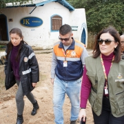 A foto mostra três pessoas caminhando em uma área externa, possivelmente um caminho de terra, com uma casa ao fundo. A mulher em primeiro plano, à direita, usa óculos de sol, um colete verde militar sobre uma blusa roxa, e calças escuras. No centro, um homem veste um colete laranja e azul da Defesa Civil e calça jeans. À esquerda, uma mulher usa uma jaqueta escura, calça cinza e sapatos pretos. Ao fundo, a casa branca tem uma placa com "JK MINERAIS". A cena parece ser de uma visita técnica ou inspeção, possivelmente relacionada a desastres naturais, dado o vestuário da Defesa Civil.
