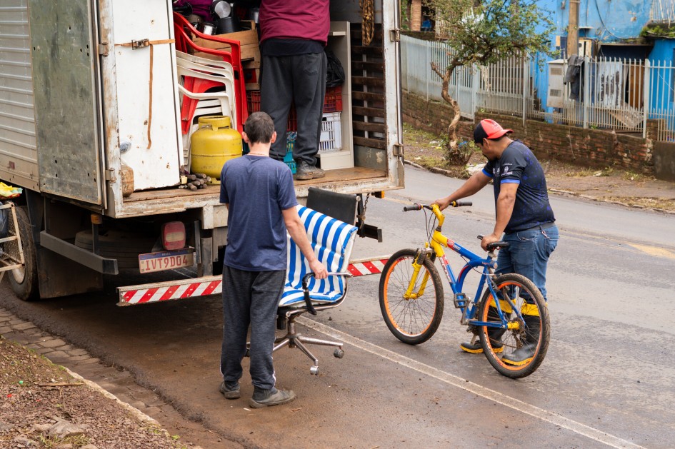 A imagem mostra três pessoas envolvidas no carregamento de itens de um caminhão. Uma pessoa está dentro do caminhão, organizando objetos. Outra pessoa, no chão ao lado do caminhão, segura uma cadeira de escritório. A terceira pessoa está segurando uma bicicleta nas cores amarela e azul.