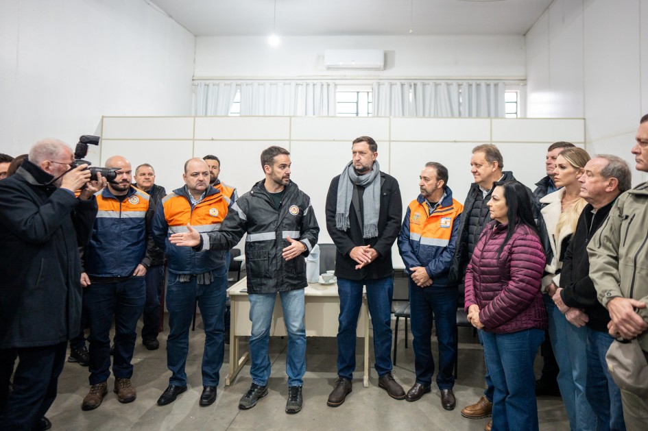 A imagem mostra um grupo de pessoas reunidas em uma sala. Algumas delas estão vestindo jaquetas da Defesa Civil. No centro da imagem, o governador está gesticulando com as mãos, falando ao grupo.
