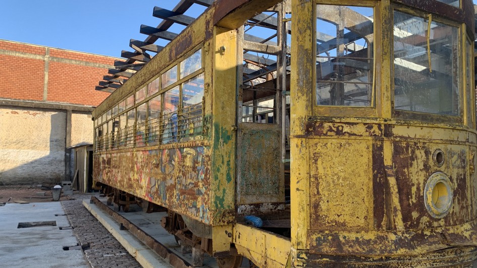 A imagem mostra um bonde antigo de cor amarela, posicionado sobre trilhos. O veículo está em estado de deterioração, com tinta descascada e ferrugem visível em várias partes da estrutura. Apesar disso, o formato original do bonde ainda é reconhecível, e a maioria das janelas permanece intacta.