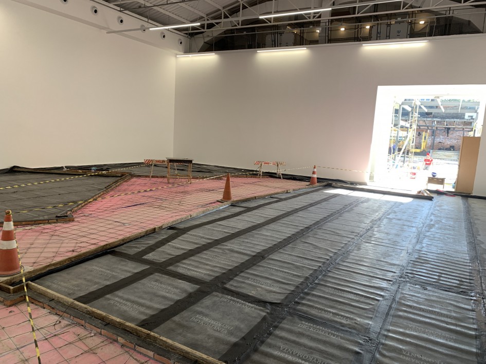 A imagem mostra um canteiro de obras interno, com um grande espaço aberto em processo de preparação. O piso está coberto com lonas plásticas pretas e estruturas metálicas. Há cones de sinalização laranja e fitas de isolamento. Ao fundo, uma grande abertura leva a uma área externa, onde também é possível observar atividades de construção em andamento. As paredes são brancas, e o teto possui vigas expostas e luminárias instaladas.
